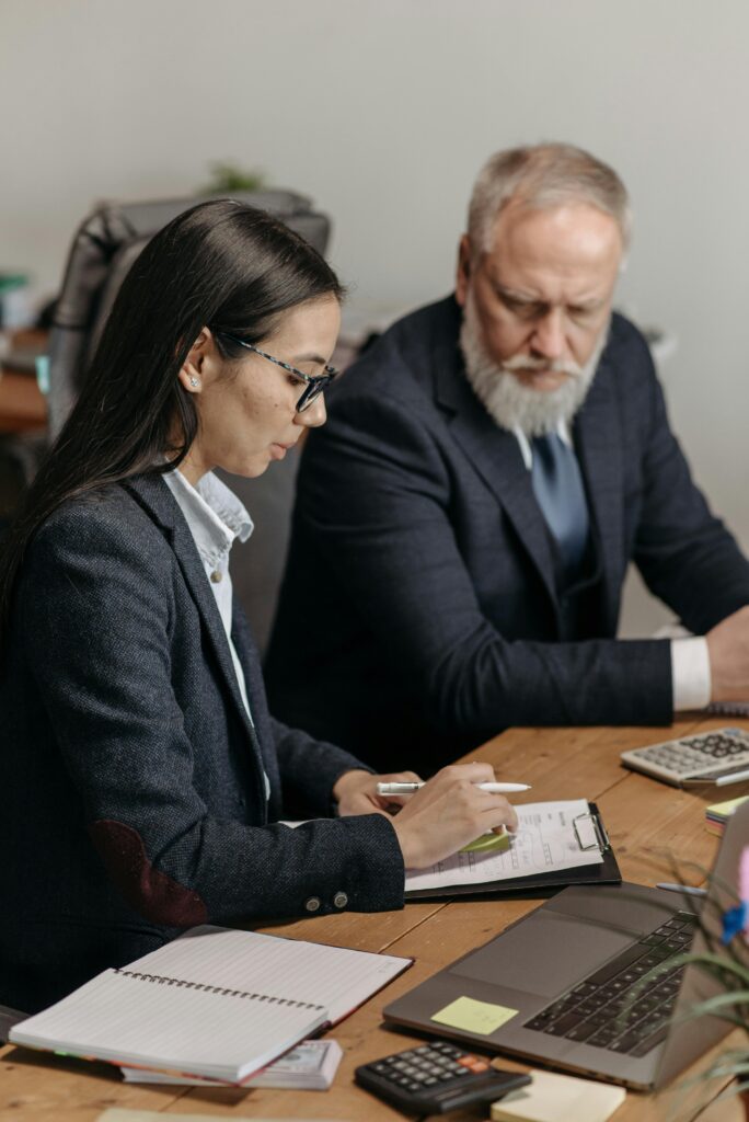 Two professionals in an office setting analyzing documents and using laptops.
